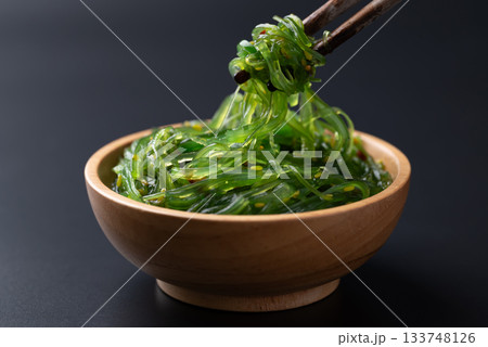 Wakame seaweed salad with sesame seed in a wooden bowl with chopsticks on black background Wakame seaweed salad with sesame seed in a wooden bowl with chopsticks on black background 133748126