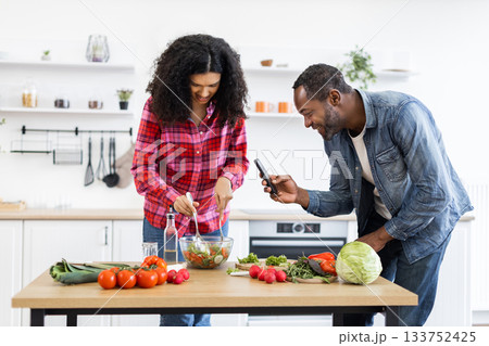 A smiling couple prepares a healthy salad in a bright, modern kitchen, with the man taking a photo of the meal. 133752425