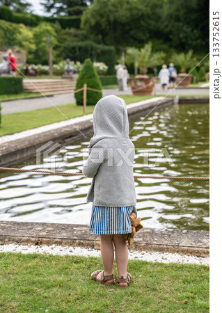 A child in a hooded sweater stands still by the pond, watching the ripples move in soft afternoon light. A child in a hooded sweater stands still by the pond, watching the ripples move in soft afternoon light. 133752615