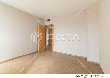 Empty room with beige walls and wooden door. The floor is tiled in light brown. Natural light enters through the open door, creating a spacious feel. 133756237
