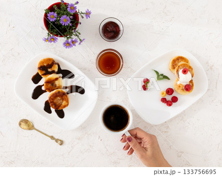 Woman with a cup of coffee and curd fritters with sour cream, berries and chocolate on the porcelain plates. Woman with a cup of coffee and curd fritters with sour cream, berries and chocolate on the porcelain plates. 133756695