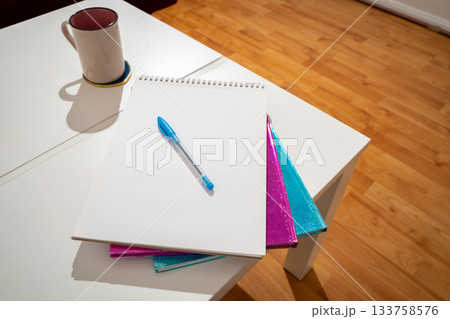 A coffee cup, a stacked notebook, and a pen resting on a blank page arranged on top of a white coffee table in soft natural light. Minimal, clean workspace setup. 133758576
