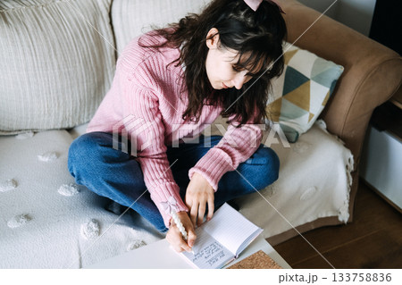 Young adult woman sits cross-legged on sofa writing goals in small notebook on coffee table. Gen Z New Year resolutions, goal setting, self improvement, personal development. Young adult woman sits cross-legged on sofa writing goals in small notebook on coffee table. Gen Z New Year resolutions, goal setting, self improvement, personal development. 133758836