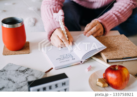 Person writes personal intentions in small notebook next to apple planning materials and red tumbler. New Year resolutions, goal setting, self improvement, healthy lifestyle. Person writes personal intentions in small notebook next to apple planning materials and red tumbler. New Year resolutions, goal setting, self improvement, healthy lifestyle. 133758921