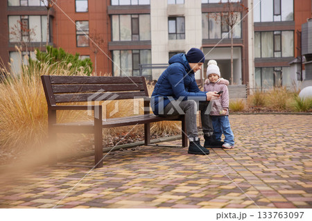 Father and child engaging with a smartphone while sitting on a bench in an urban setting 133763097