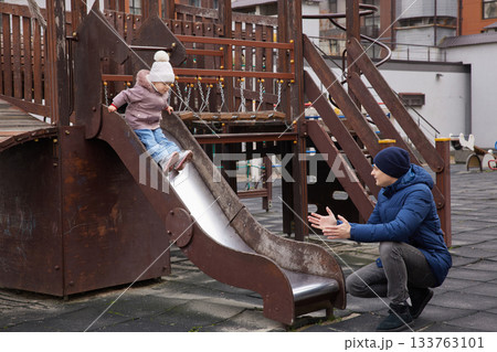 Father catching his toddler daughter sliding down a playground slide 133763101