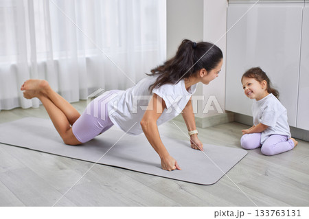 Mother doing push-ups on a mat, daughter smiling and watching Mother doing push-ups on a mat, daughter smiling and watching 133763131