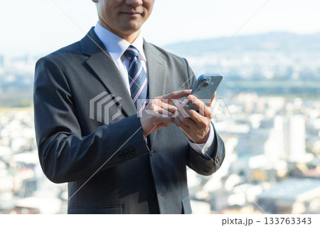 町を背景にスマホを持つスーツを着たミドル男性ビジネスマン 133763343