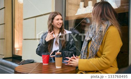 Young women talking over coffee at outdoor cafe table in cozy autumn street Young women talking over coffee at outdoor cafe table in cozy autumn street 133768495