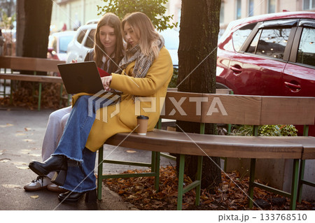Outdoor study session on bench with two young women using laptop and takeaway coffee 133768510