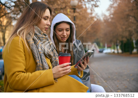Two young women with coffee outdoors checking smartphone in autumn scenery Two young women with coffee outdoors checking smartphone in autumn scenery 133768512