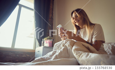 Smiling young woman using smartphone in cozy bedroom morning light 133768530
