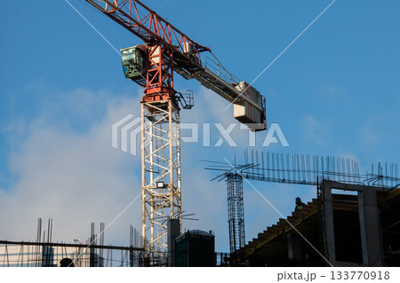 Control Cabin and Counterweight of Red Tower Crane at Construction Site. Monolithic Box with Window Holes and Rebar outlets against Blue Sky background. Control Cabin and Counterweight of Red Tower Crane at Construction Site. Monolithic Box with Window Holes and Rebar outlets against Blue Sky background. 133770918