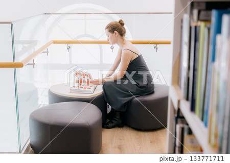 woman in library is reading book sitting on wide chair. lady in black dress flips through pages. woman in library is reading book sitting on wide chair. lady in black dress flips through pages. 133771711