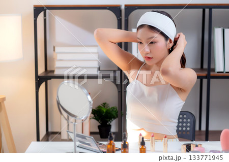 Young asian woman wearing headband and tying hair while preparing skincare looking mirror. Young asian woman wearing headband and tying hair while preparing skincare looking mirror. 133771945