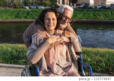 Senior woman in wheelchair walking with caregiver old man on road in park. Elderly family couple man supporting embracing woman in chair for people with disability outdoor. Rehabilitation 133774647