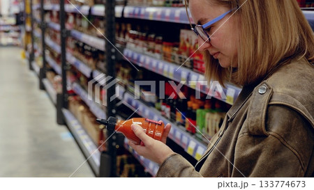 Customer reading label of red hot sauce bottle while shopping groceries in supermarket, standing in front of shelf with various food products 133774673