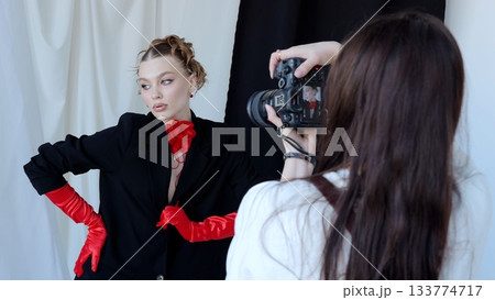 Fashion photographer taking pictures of a beautiful model wearing red gloves and a red flower choker, posing in a studio with black and white background 133774717