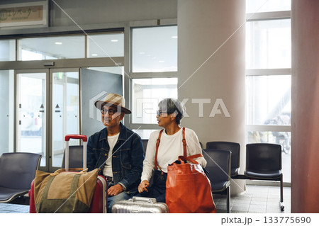 待合室でスーツケースの隣に座る中高年の旅行客カップル 133775690