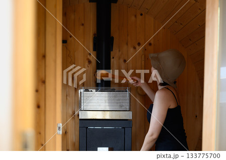 サウナストーブに水をかける女性、樽型サウナ内の光景 133775700