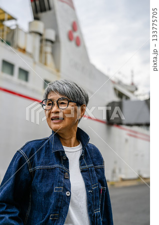 デニムジャケットを着たグレーヘアの年配女性、フェリーを背景に港で微笑む デニムジャケットを着たグレーヘアの年配女性、フェリーを背景に港で微笑む 133775705