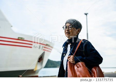 港に停泊するフェリーを背景に立つ、旅行かばんを持った高齢の日本人女性 133775735