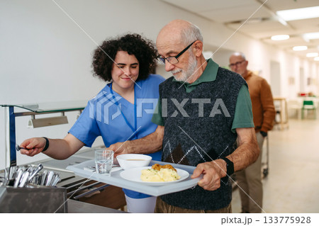 Senior man having lunch with supportive caregiver in community center cafeteria. 133775928