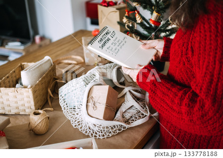 Young adult woman in red sweater reads handwritten Christmas shopping list on notebook next to small decorated tree. Christmas checklist, organized celebration, planning ahead, mindful gifting. Young adult woman in red sweater reads handwritten Christmas shopping list on notebook next to small decorated tree. Christmas checklist, organized celebration, planning ahead, mindful gifting. 133778188