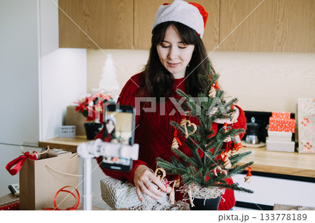 Young adult woman wearing Santa hat records video on smartphone while decorating small Christmas tree. Xmas holiday influencer, Christmas vlog, festive content, seasonal social media. Young adult woman wearing Santa hat records video on smartphone while decorating small Christmas tree. Xmas holiday influencer, Christmas vlog, festive content, seasonal social media. 133778189