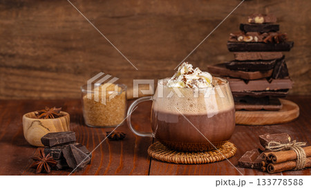 Hot chocolate with whipped cream served in a glass mug on a rustic wooden table surrounded by dark chocolate pieces, cinnamon sticks, star anise and brown sugar, creating a warm cozy winter atmosphere 133778588
