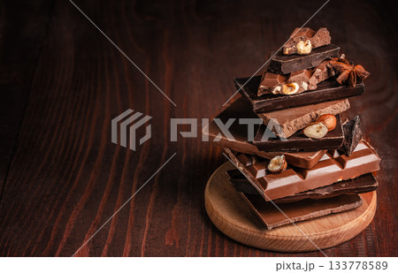 Assortment of stacked chocolate bars with nuts and spices on a wooden board placed on a dark rustic wooden background copy space, creating a warm and rich confectionery atmosphere for food 133778589