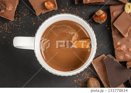 Hot chocolate with cinnamon in a white cup surrounded by chocolate pieces, hazelnuts and spices on a dark textured background, top view flat lay copy space for cozy winter dessert concepts. Hot chocolate with cinnamon in a white cup surrounded by chocolate pieces, hazelnuts and spices on a dark textured background, top view flat lay copy space for cozy winter dessert concepts. 133778634