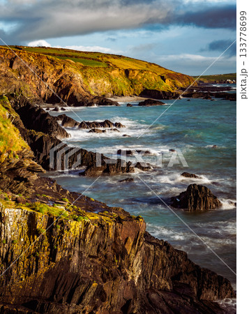 A breathtaking view of the rugged Irish coastline. Ocean waves crash against the rocks below the cliffs. The sky is blue with scattered clouds, showing a beautiful landscape. A breathtaking view of the rugged Irish coastline. Ocean waves crash against the rocks below the cliffs. The sky is blue with scattered clouds, showing a beautiful landscape. 133778699