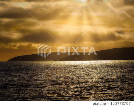 Golden sunlight filters through clouds and illuminates the ocean surface. Dark land masses are visible in the distance against the warm, colorful sky. Golden sunlight filters through clouds and illuminates the ocean surface. Dark land masses are visible in the distance against the warm, colorful sky. 133778707
