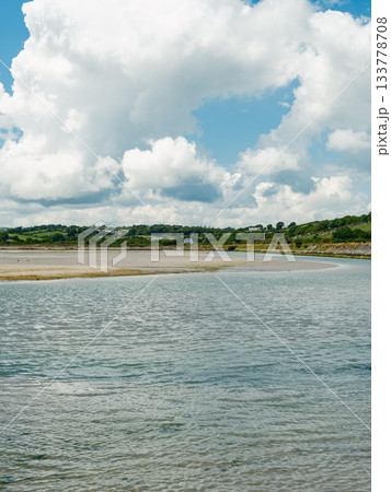 A picturesque landscape featuring shallow water, a sandy shoreline, green hills, and a beautiful sky filled with clouds. 133778708