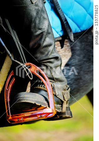 close-up of a brightly colored stirrup, with a rider's booted and cuisse-clad foot and a dressage whip in it. Photographed on a sunny day during equestrian training close-up of a brightly colored stirrup, with a rider's booted and cuisse-clad foot and a dressage whip in it. Photographed on a sunny day during equestrian training 133778825