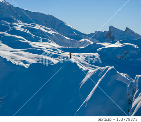 Aerial photo of a ski mountaineer standing on a snowy alpine ridge in Austria. Winter landscape, backcountry adventure, clear blue sky with sunshine and solitude in breathtaking mountain scenery. 133778875