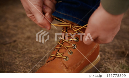 A Close-Up Image of Hands Tying Laces on a Brown Boot, Highlighting the Texture and Detail of Both the Footwear and the Surrounding Natural Environment 133778899