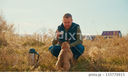 A Person Enjoys a Warm Beverage Outdoors While Spending Quality Time with Their Loyal Dog in a Sunlit Field Surrounded by Tall Grass and Nature's Beautiful Scenery 133778915