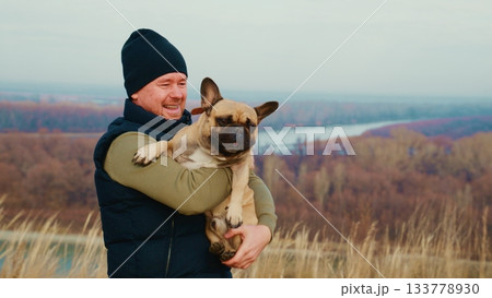A Happy Man Embraces His French Bulldog in a Scenic Outdoor Landscape, Showcasing the Bond Between Pets and Their Owners in a Majestic Natural Setting A Happy Man Embraces His French Bulldog in a Scenic Outdoor Landscape, Showcasing the Bond Between Pets and Their Owners in a Majestic Natural Setting 133778930