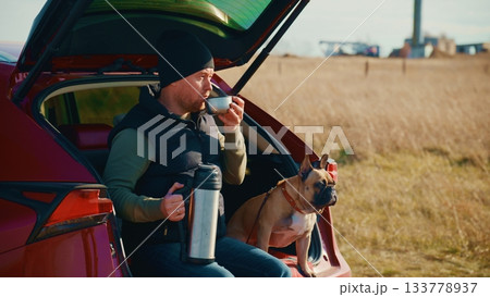 A man enjoying a cup of coffee while seated in the trunk of a car, accompanied by a French Bulldog, amidst a scenic landscape with fields and a clear blue sky in the background. 133778937