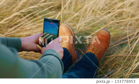 Capturing Nature's Beauty: A Person Sitting in a Field of Grass While Taking a Photo with a Smartphone, Showcasing the Serenity of the Outdoor Environment and Relaxation 133778953