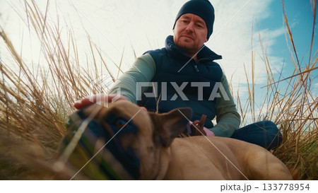 A Relaxing Moment in Nature as a Man Enjoys Time with His French Bulldog in a Serene Field Surrounded by Tall Grass and a Beautiful Sky 133778954