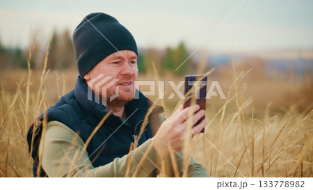 A man dressed in warm clothing sits in a field of tall grass while using a smartphone, enjoying a moment of connection with nature and technology in a tranquil outdoor setting. A man dressed in warm clothing sits in a field of tall grass while using a smartphone, enjoying a moment of connection with nature and technology in a tranquil outdoor setting. 133778982