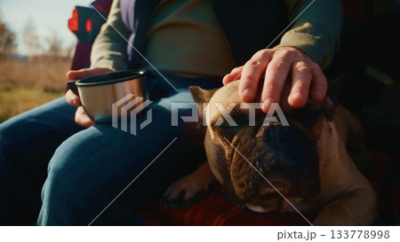 A Peaceful Moment in Nature: A Man Enjoys a Hot Drink While Petting His Content French Bulldog in a Tranquil Outdoor Setting Surrounded by Scenic Views and Warm Sunlight A Peaceful Moment in Nature: A Man Enjoys a Hot Drink While Petting His Content French Bulldog in a Tranquil Outdoor Setting Surrounded by Scenic Views and Warm Sunlight 133778998