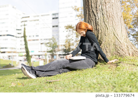 Young woman reading book leaning against tree in park 133779674