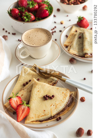 Crepes with chocolate hazelnut spread, strawberries and hazelnuts on a white wooden table. 133780292