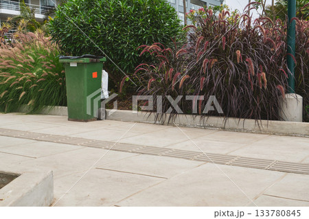 Green trash can standing on urban sidewalk surrounded by plants Green trash can standing on urban sidewalk surrounded by plants 133780845