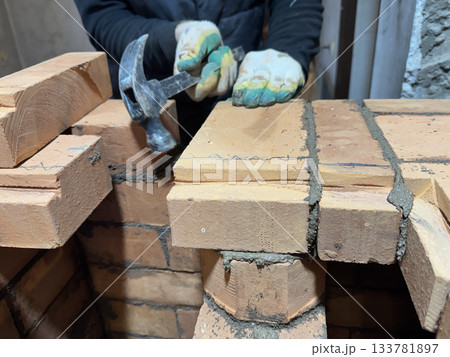 A stove fitter adjusts a laid brick using a hammer. Brickwork of a heating stove. Channels for dividing and passing smoke. Close-up. 133781897