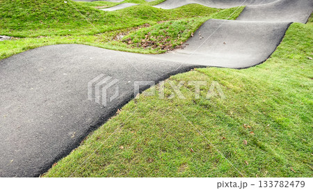 Curved asphalt pump track for cycling and wheeled sports, surrounded by vibrant green grass in a modern park 133782479